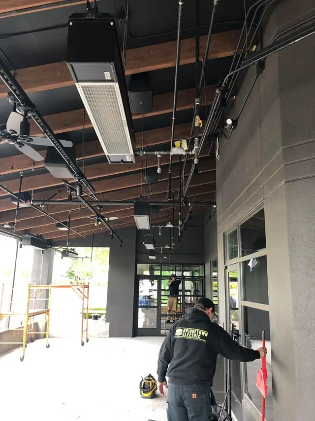 Bridgetown Electric technician in a branded jacket wiring overhead heaters on a commercial patio